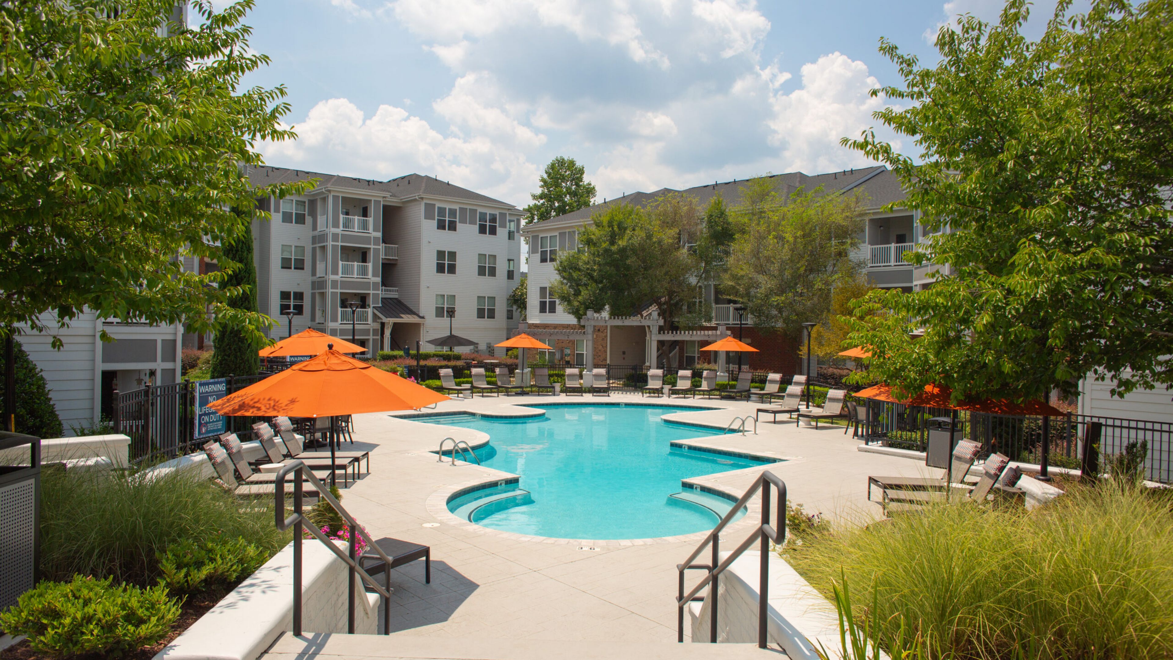 Hawthorne Davis Park outdoor area of an apartment complex. The scene features several lounge chairs around a shaded pergola with trees and shrubbery providing a backdrop. Multiple apartment buildings with white exteriors surround the area under a bright blue sky.